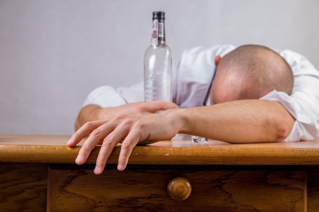 image shows a man slumped over a desk drunk with an empty bottle of alcohol in front of him